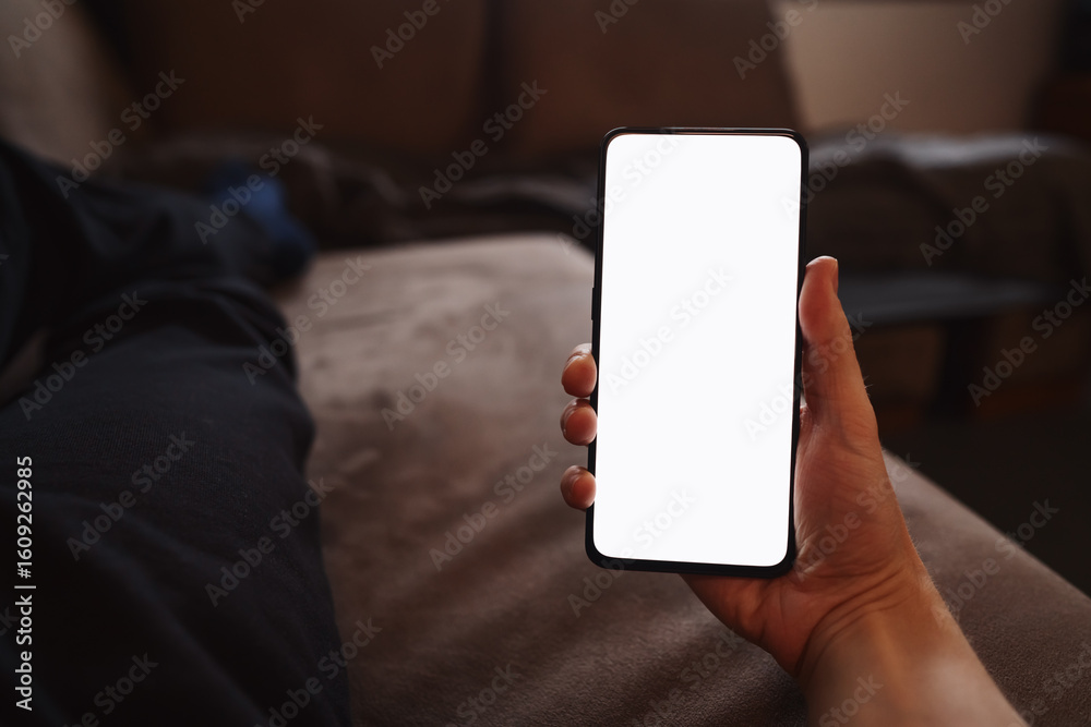 Man hold smartphone with white screen in his hand lying on a couch in home environment Man hold smartphone with white screen in his hand lying on a couch in home environment