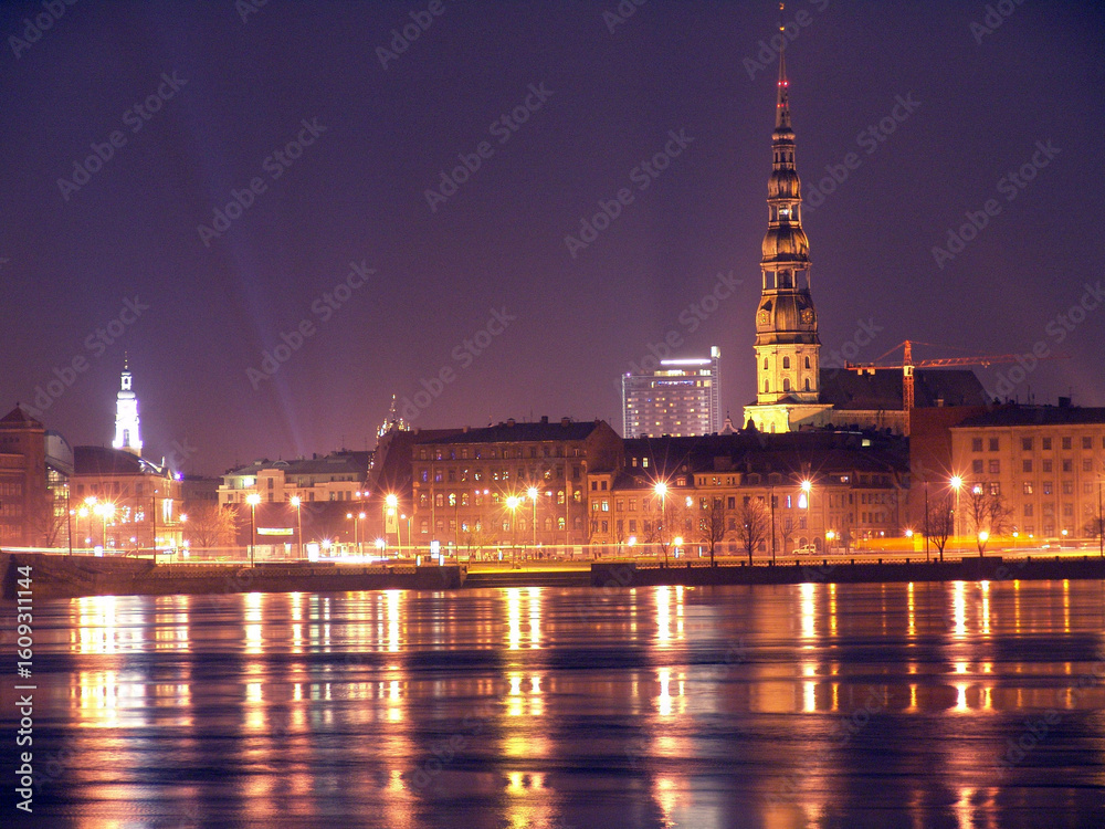 Obraz premium Night cityscape of Riga, Latvia, featuring illuminated buildings, St. Peter's Church tower, and shimmering reflections on the Daugava River.