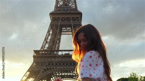 Peaceful moment of a young woman standing by the Eiffel Tower during golden hour eyes closed and smiling in the warm Parisian light