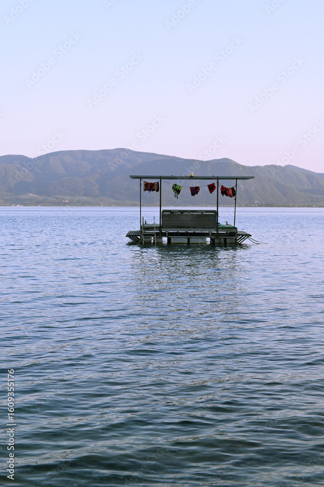 Fototapeta premium A small floating platform with colorful flags stands still on a calm sea, surrounded by peaceful blue water and distant mountain hills. The clear sky and tranquil setting evoke a sense of serenity, so