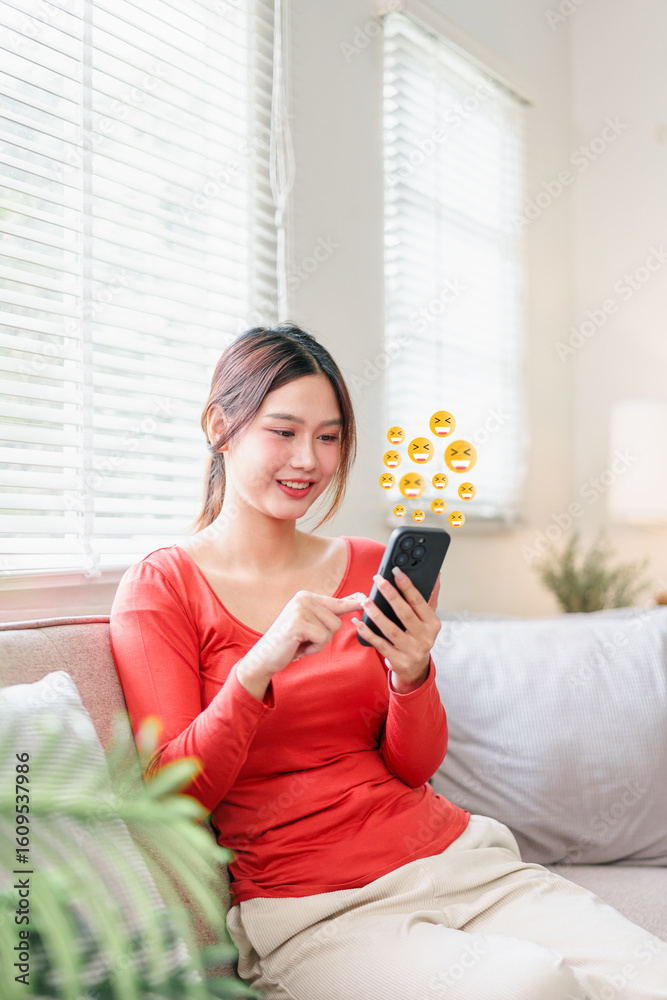 Young Asian woman in red shirt sitting on sofa, smiling and using smartphone with floating emoji icons, bright living room, cheerful mood Young Asian woman in red shirt sitting on sofa, smiling and using smartphone with floating emoji icons, bright living room, cheerful mood