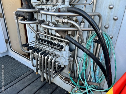Hydraulic control panel on fishing boat, with multiple stainless steel lines, valves, and connections alongside coiled green rope on a vessel deck