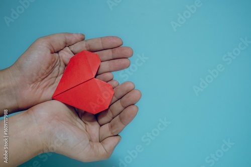 Vibrant red origami heart gently held in cupped hands against a calming blue background with copy space. Conveys love, care, giving, compassion, and heartfelt gestures.