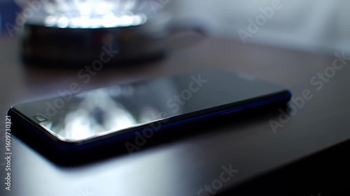 A blue-edged phone lies on a dark wooden surface, reflecting blurred shapes on its screen