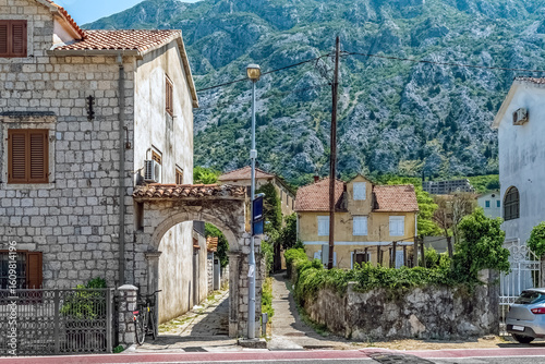 Charming stone houses and an old archway in Kotor, Montenegro, with a stunning backdrop of green mountains. This picturesque view showcases the peaceful residential streets of the historic town