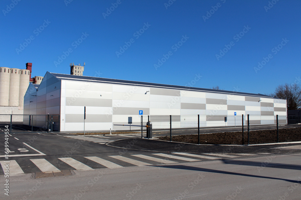 A new white-paneled warehouse sits within a gated, empty parking lot, with industrial silos and older factory buildings rising in the distant blue sky A new white-paneled warehouse sits within a gated, empty parking lot, with industrial silos and older factory buildings rising in the distant blue sky