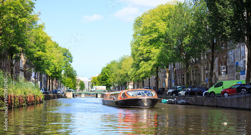 Low Angle view of Amsterdam Canal with an orange passenger vessel sailing forward along a tree-lined road