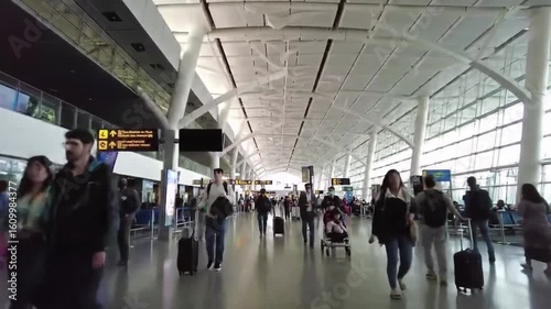 Busy airport terminal filled with travelers walking towards their gates, with digital flight information displays and large windows letting in natural light in the background