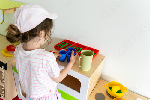 Fotografi child arranging toy cups and a pitcher on a wooden toy kitchen counter, with dishes and utensils