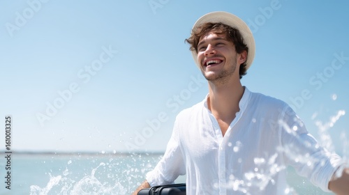 A happy young man enjoys a sunny day by the sea. He splashes water while wearing a stylish white shirt and a straw hat. This image captures joy and relaxation. 