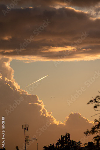 An airplane flies out of a bank of dramatic, sunlit clouds at sunset, leaving a long white contrail across the golden evening sky.