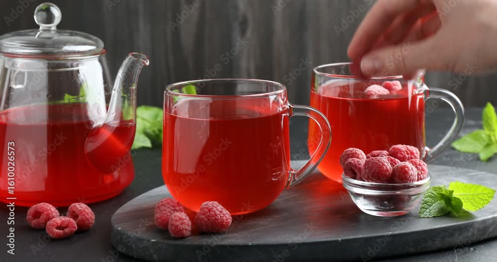 Woman adding fresh raspberries into glass cup with tea at black table, closeup