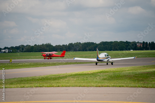 Two small light aircraft taxiing on airport runway on cloudy day, general aviation planes preparing for takeoff on green airfield with grey tarmac underneath