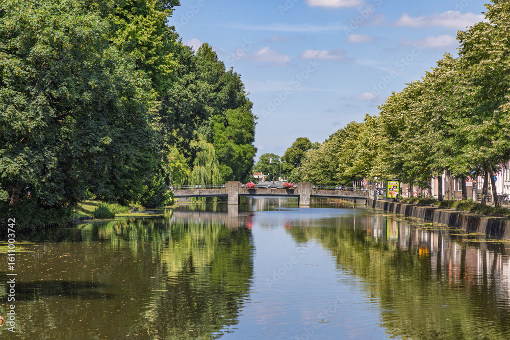 Fototapeta premium A wide view of a serene canal in Gouda, Netherlands, flanked by dense green trees and featuring a bridge in the distance, with calm water. Gouda, Netherlands, 29 June 2025.
