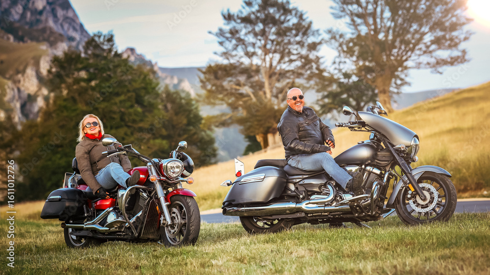 Obraz premium Happy mature couple enjoying a motorcycle road trip in nature, smiling and relaxing with their cruisers parked on a scenic countryside background.