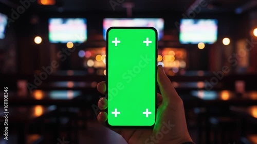 Hand holding green screen phone in bar with blurred tvs and lights in the background at nighttime