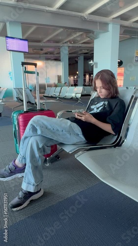Young teenage girl sitting in airport lounge, focused on smartphone while waiting for flight, with colorful luggage beside her and modern seating in a bright, spacious terminal environment