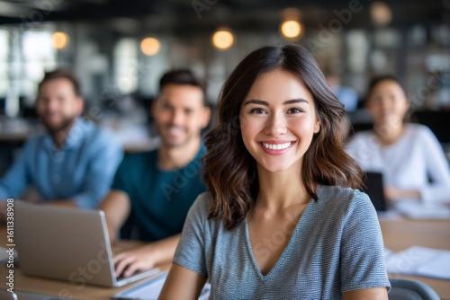 A confident young woman smiles at the camera while sitting in a classroom with peers working on laptops in the background.