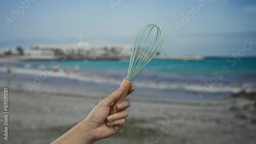 Hand of a man holding a whisk on a beach with the sea in the background, capturing a unique seaside scene.