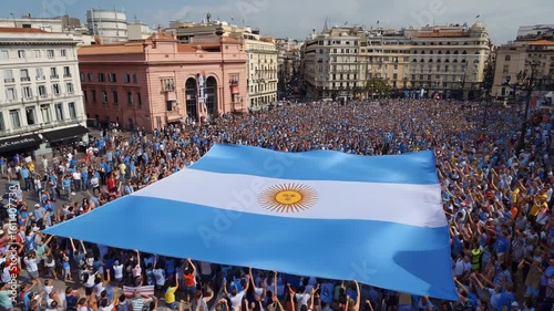 Massive Argentine flag waves over jubilant crowd celebrating victory in vibrant city square, a powerful display of national pride and collective euphoria during a momentous event.