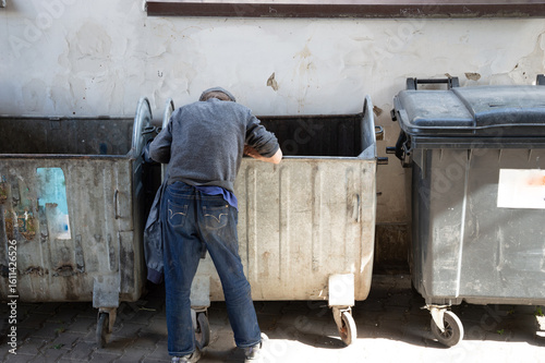 Individual rummages through dumpsters in a city alley, searching for food or discarded items in the sunlight
