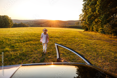 Woman walking away from car into summer sunset field. Concept of freedom, escapism and joy of travel