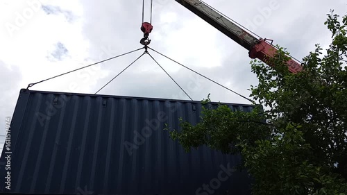 A construction crane with a fully extended boom lifts a large metal container into the air. Captured in an industrial setting under natural daylight. Ideal for themes of heavy lifting, logistics