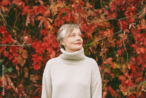 Autumn portrait of happy mature woman wearing beige pullover or sweater, posing outdoors with red ivy red leaves background