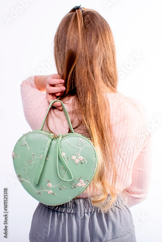 Green heart-shaped handbag in the girl's hand. Holds the bag behind her back
