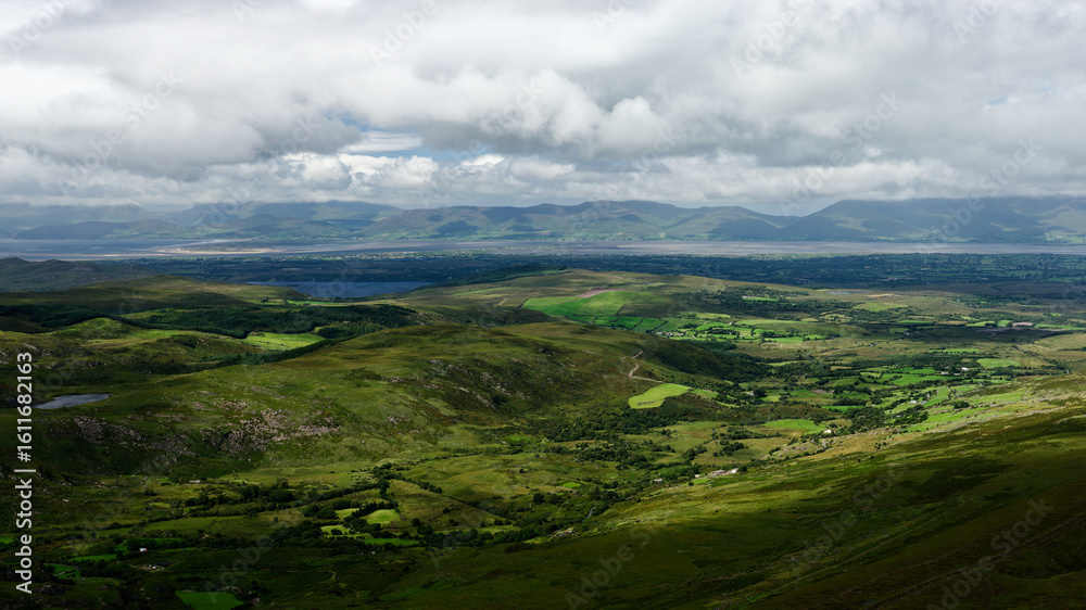 Obraz premium Rolling green valleys with interspersed forests and open pasture lead to distant mountains under a cloudy sky, taken from Coomloughra Horseshoe scenic landscape in Ireland.