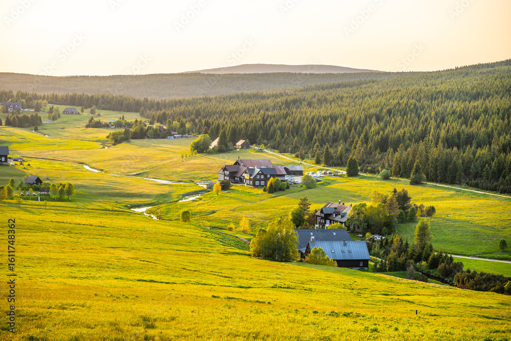 Naklejka premium A serene evening unfolds in Jizerka Village, nestled in the Jizera Mountains. The golden sunlight casts a warm glow over lush green fields and charming houses, creating a peaceful atmosphere.