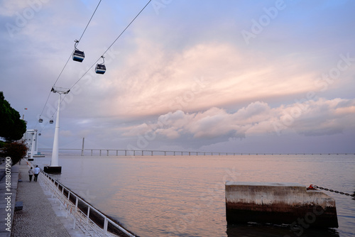 Fotografie A scenic view of a waterfront promenade with a cable car system overhead, trees lining the walkway, and a calm sea under a cloudy sky