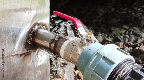 Water flows steadily from a metal valve tap with a red handle. The scene highlights utility, plumbing, and industrial simplicity. Ideal for themes of water supply, maintenance, or minimalism.