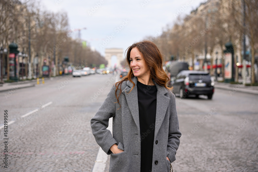 Fototapeta premium Young woman in gray coat walking along Champs Elysees in Paris with Arc de Triomphe visible in the background
