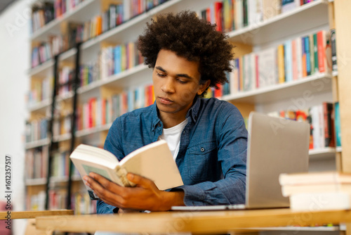 Focused Latin male student immersing in exam preparation, reading book, sitting in university library after lectures and classes