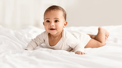 Childcare. Portrait of cute little African American baby wearing bodysuit lying on the white blanket at home. Black infant child crawling on bed in the bedroom Selective focus, blurred background
