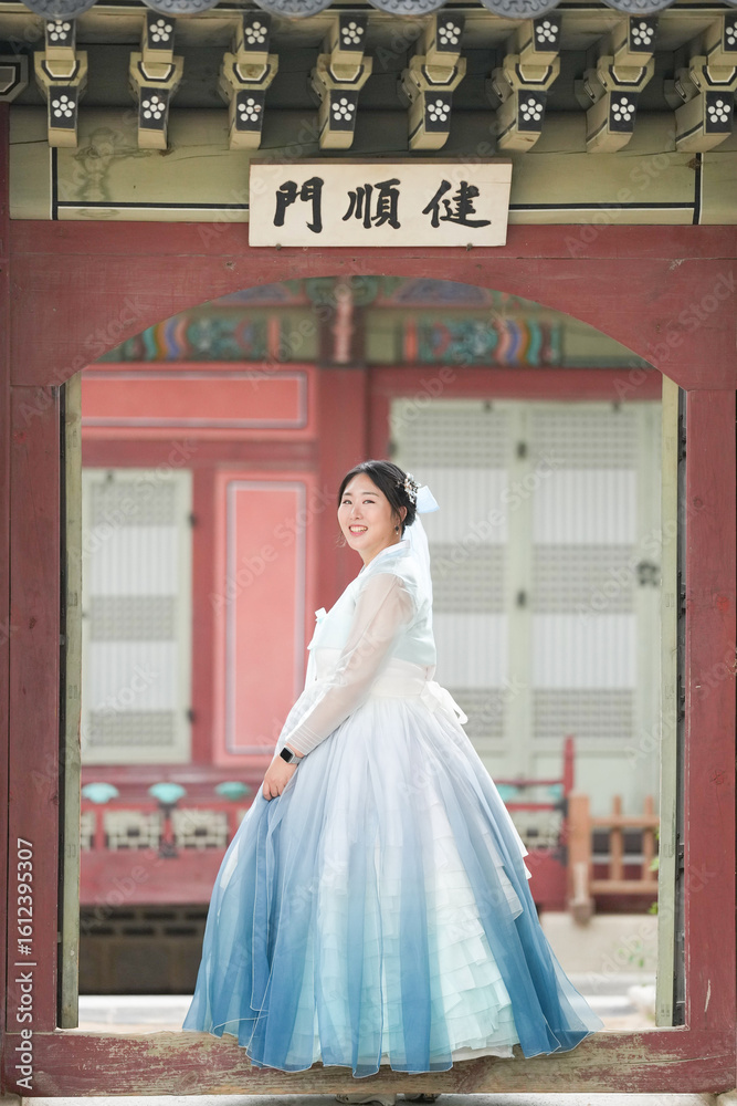 Fototapeta premium A Korean woman in her twenties stands gracefully before the brick gate of Gyeongbokgung Palace, wearing a sky-blue hanbok and admiring the historic architecture under a clear summer sky. Seoul, June.