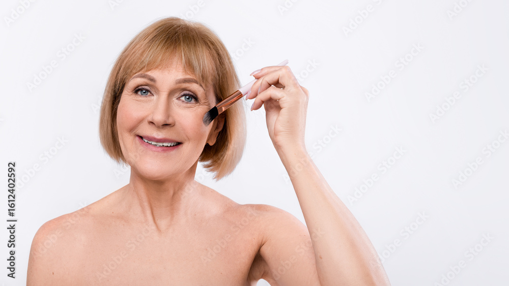 Beautiful elderly lady applying blush with brush, posing half naked on light background, copy space Beautiful elderly lady applying blush with brush, posing half naked on light background, copy space