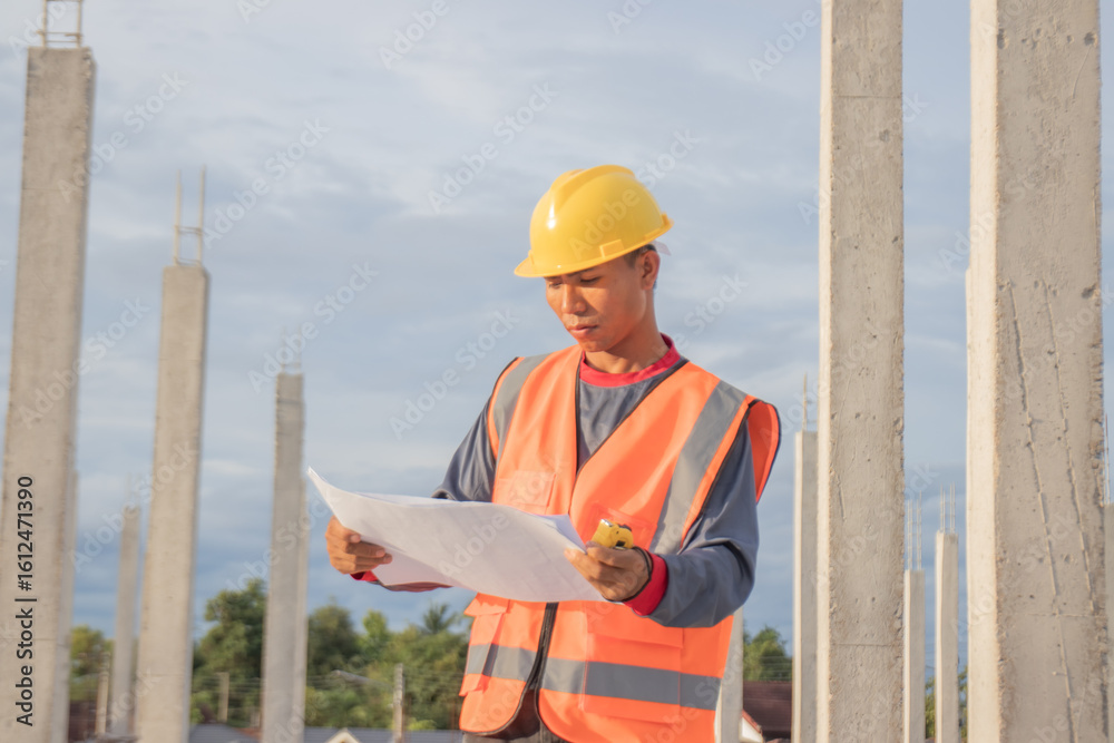 Fototapeta premium Male engineer in hard hat and safety vest examines blueprint indoors at construction site; planning infrastructure project with expertise, teamwork, and efficiency.