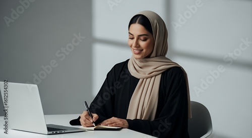 A focused young woman in a hijab works diligently on her laptop and takes notes in a notebook, showcasing modern professionalism and Islamic attire.