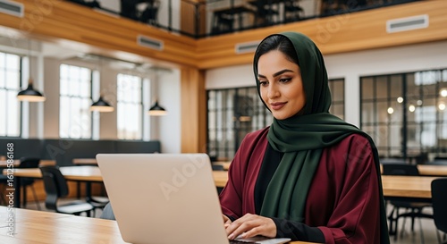 A young woman in a hijab works on her laptop in a modern, open-plan space.