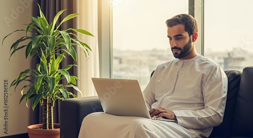 A young man in traditional Middle Eastern attire works on his laptop, comfortably seated on a couch near a large houseplant in a bright, sunlit room.