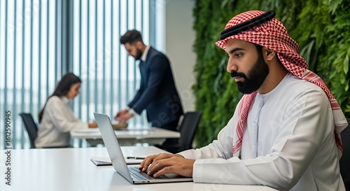 A businessman in traditional Middle Eastern attire works on his laptop in a modern, collaborative space.