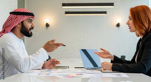 A man in traditional Middle Eastern attire and a woman in business attire engage in a serious discussion over a laptop during a meeting.