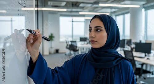 A young woman in a hijab thoughtfully writes equations on a whiteboard during a brainstorming session in a modern .