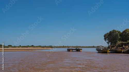 Exotic ferry crossing on the African red-brown river. Double boats with decking between decks and canopies. Green vegetation on sandy shores. The blue sky. Copy space. Madagascar. Tsiribihina  river