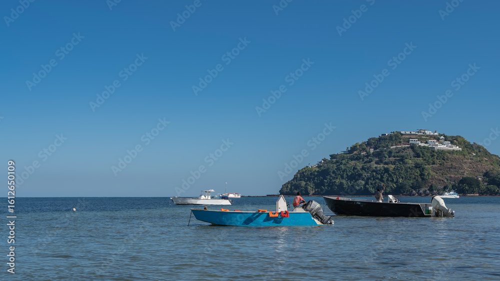 Obraz premium Motor boats are anchored in the blue ocean. White buildings on a green hill. Clear azure sky. Copy space. Madagascar. Nosy Be 