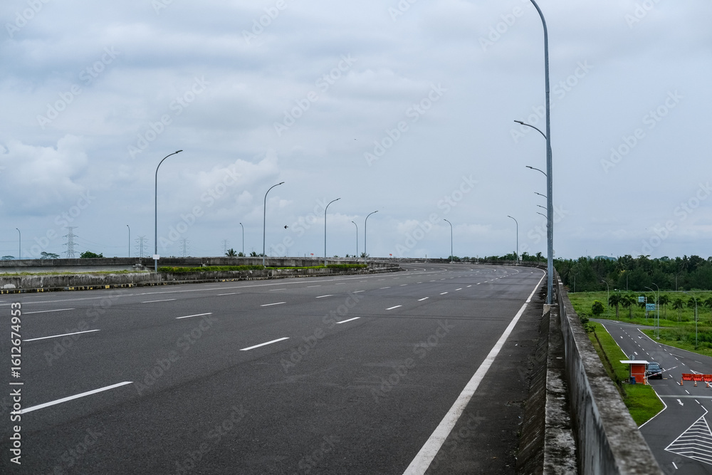 Fototapeta premium Empty road stretches over a bridge under cloudy skies, surrounded by green fields and minimal traffic, offering a quiet scene of infrastructure and open space. Road in pandemic time.