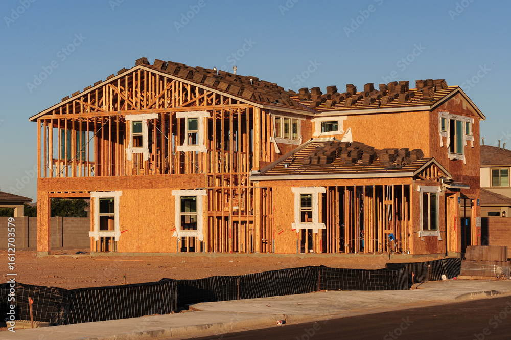 Fototapeta premium A wood-framed two-story house is under construction with roof tiles staged and framing exposed, representing active residential development in a suburban neighborhood
