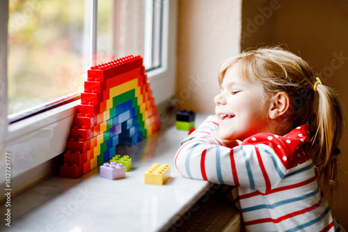 Cute little toddler girl by window create rainbow with colorful plastic blocks during pandemic coronavirus quarantine. Children made and paint rainbows around the world as sign.